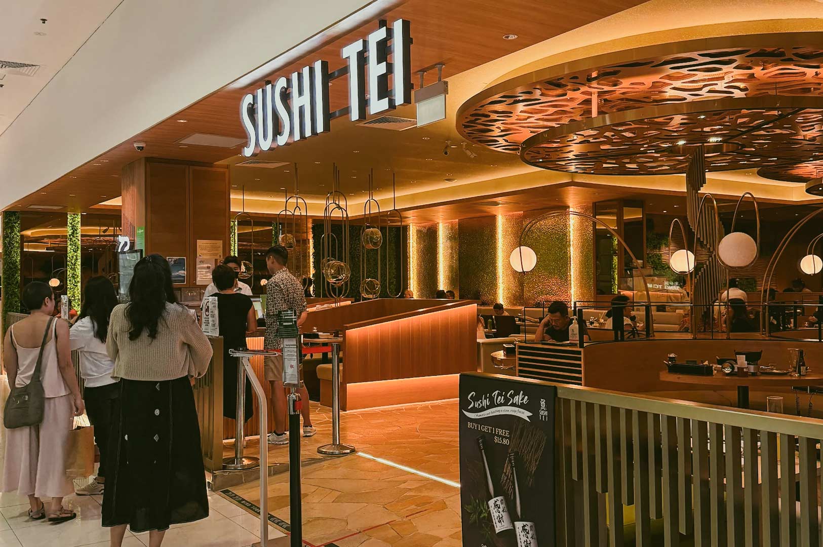 Sushi Tei Japanese restaurant entrance in shopping mall with promotional sake sign, stylish design, and customer queue waiting to dine.
