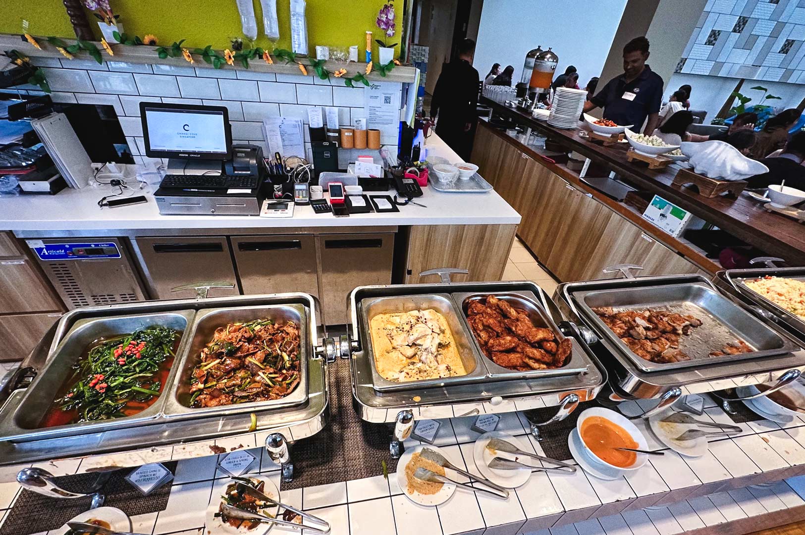 Buffet counter at The White Olive displaying a variety of hot dishes in stainless steel chafing trays, including vegetables, stir‑fried meats, roasted chicken, and creamy pasta, with sauces and plates arranged in front.”