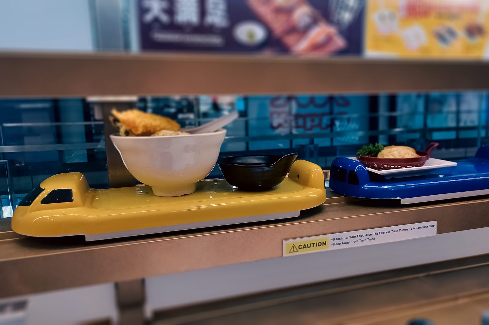 Automated train-style food delivery system at Genki Sushi, carrying bowls of noodles and Japanese dishes.