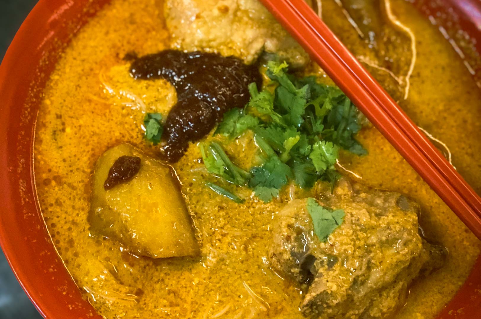 Close-up shot of curry mee in red bowl with coconut broth, chicken, potato, egg, sambal, and cilantro garnish.