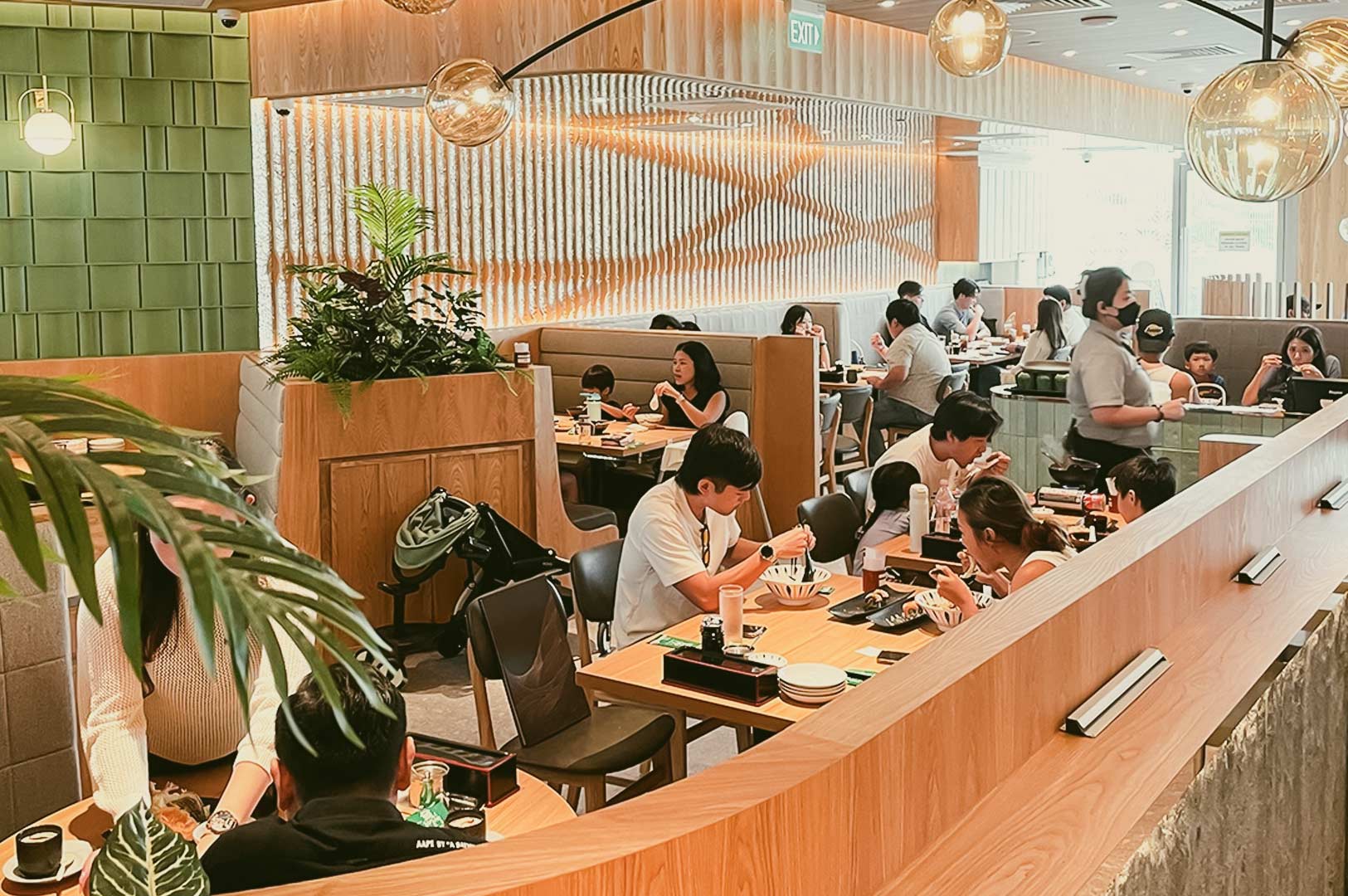 Busy Sushi Tei restaurant scene with families and groups enjoying meals, warm lighting, green wall tiles, and decorative spherical fixtures.