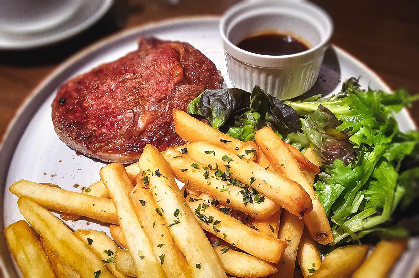 Plate of steak served with golden fries, mixed green salad, and a small cup of brown sauce.