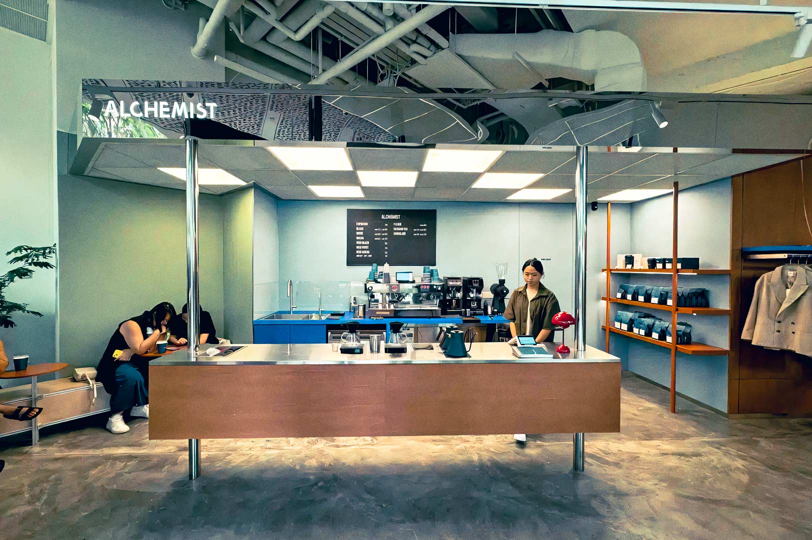 Interior of Alchemist café featuring a minimalist counter, espresso machines, wall shelving with products, and a modern industrial ceiling design.