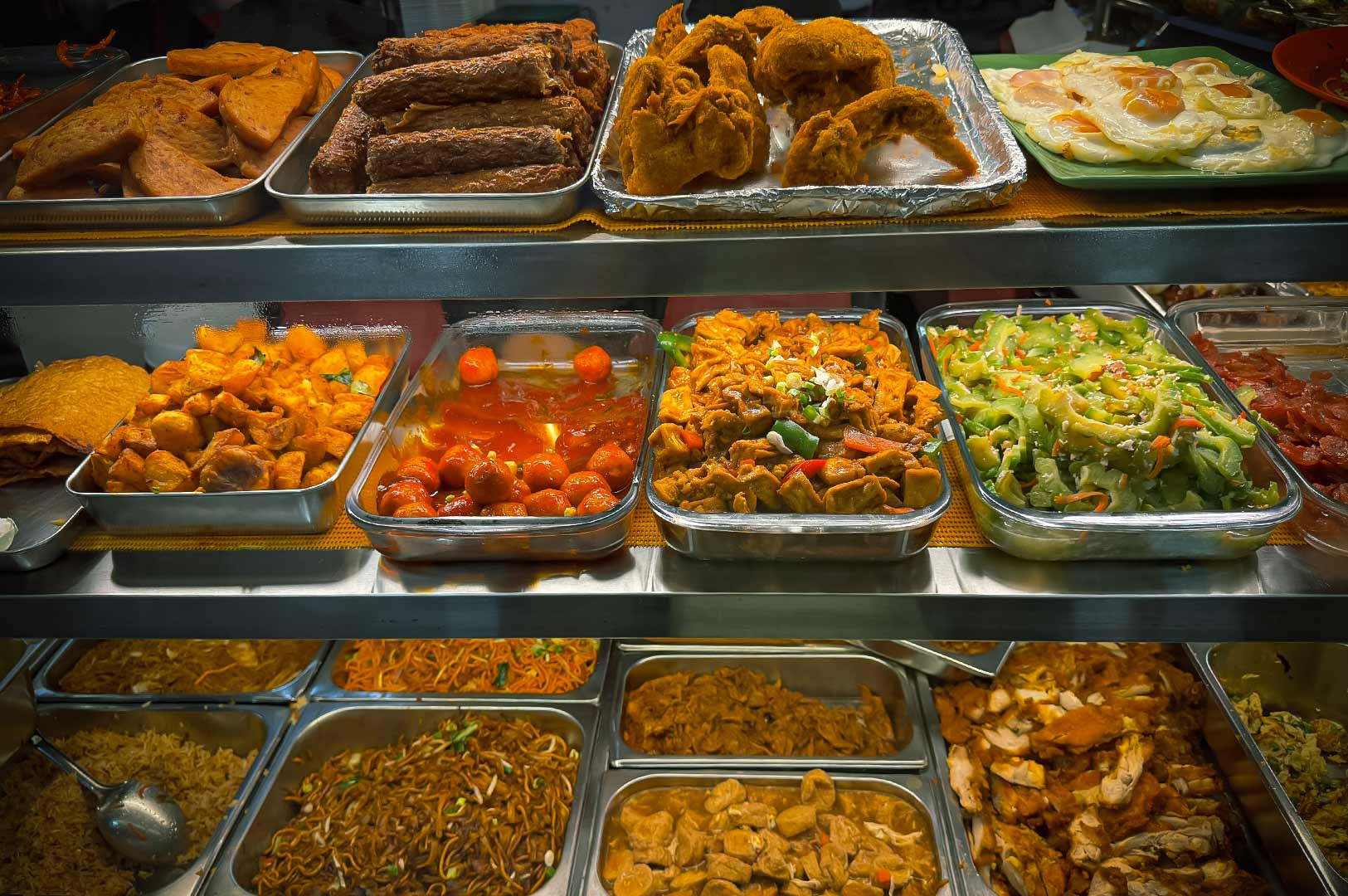 Food court counter showcasing diverse Asian dishes including fried items, vegetables, noodles, rice, and stir-fried specialties.