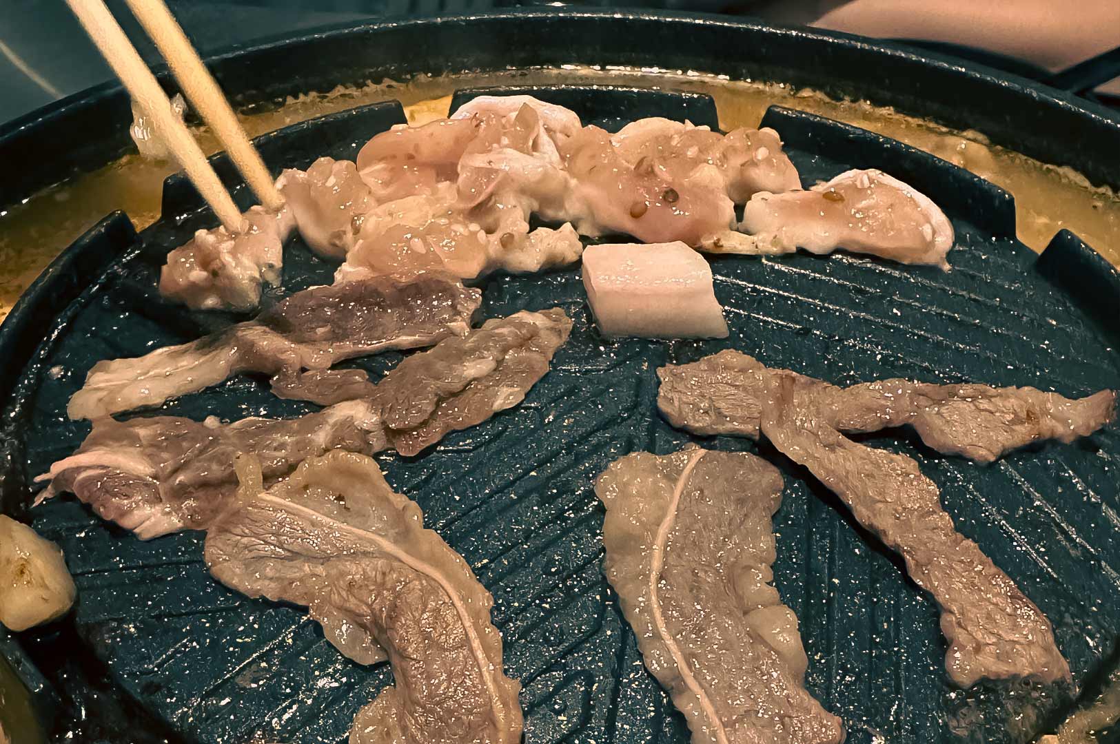 Slices of pork and chicken grilling on a Mookata dome pan with chopsticks cooking the meat.