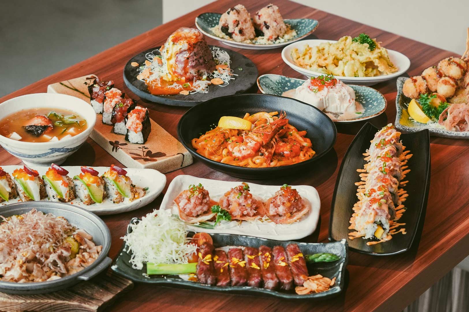 Assorted Japanese dishes including sushi rolls, sashimi, grilled meat, tempura, and seafood pasta on wooden table.