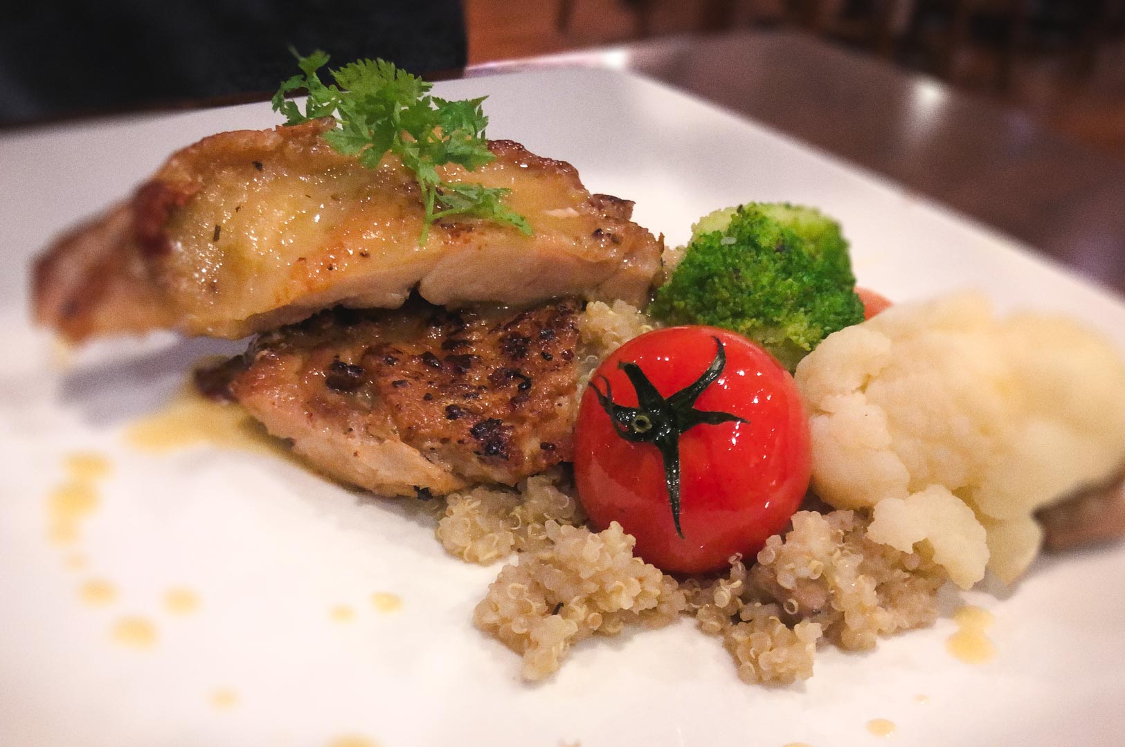 Plate of grilled chicken served on quinoa with a roasted cherry tomato, broccoli, and cauliflower.