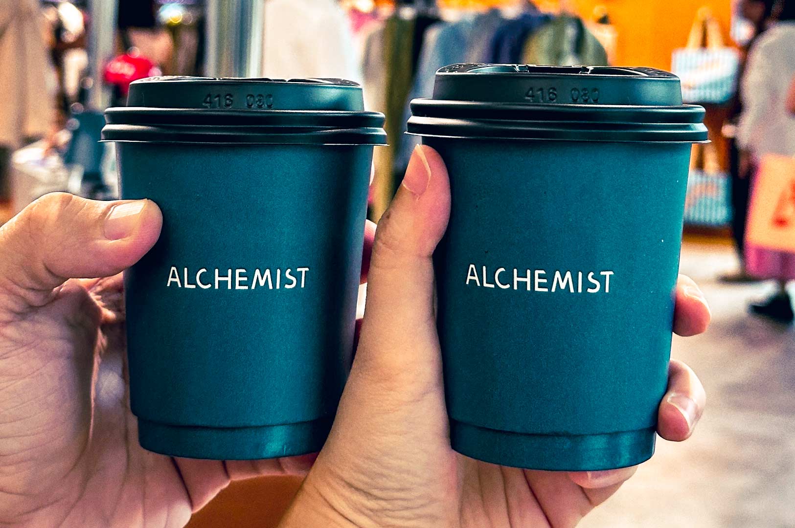 Two hands holding takeaway coffee cups with the Alchemist logo against a café background.