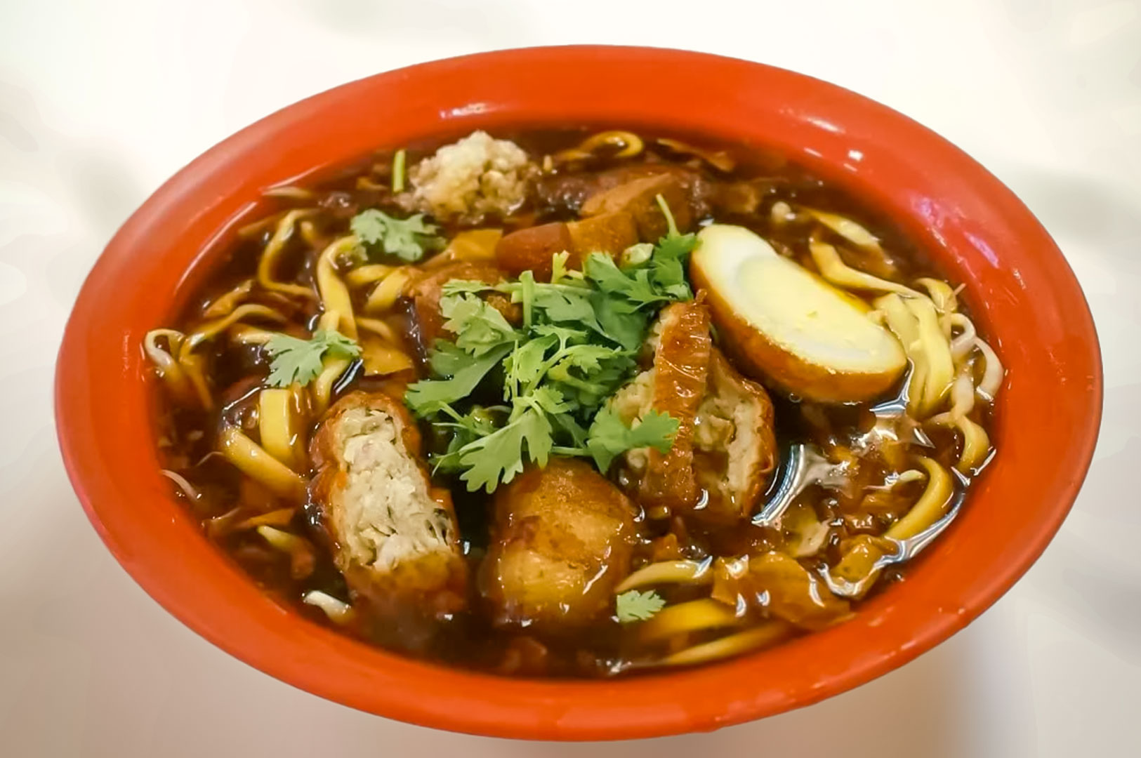 Savory Lor Mee served in red bowl, featuring wheat noodles in rich dark broth topped with braised meat, egg, fried rolls, and fresh cilantro.