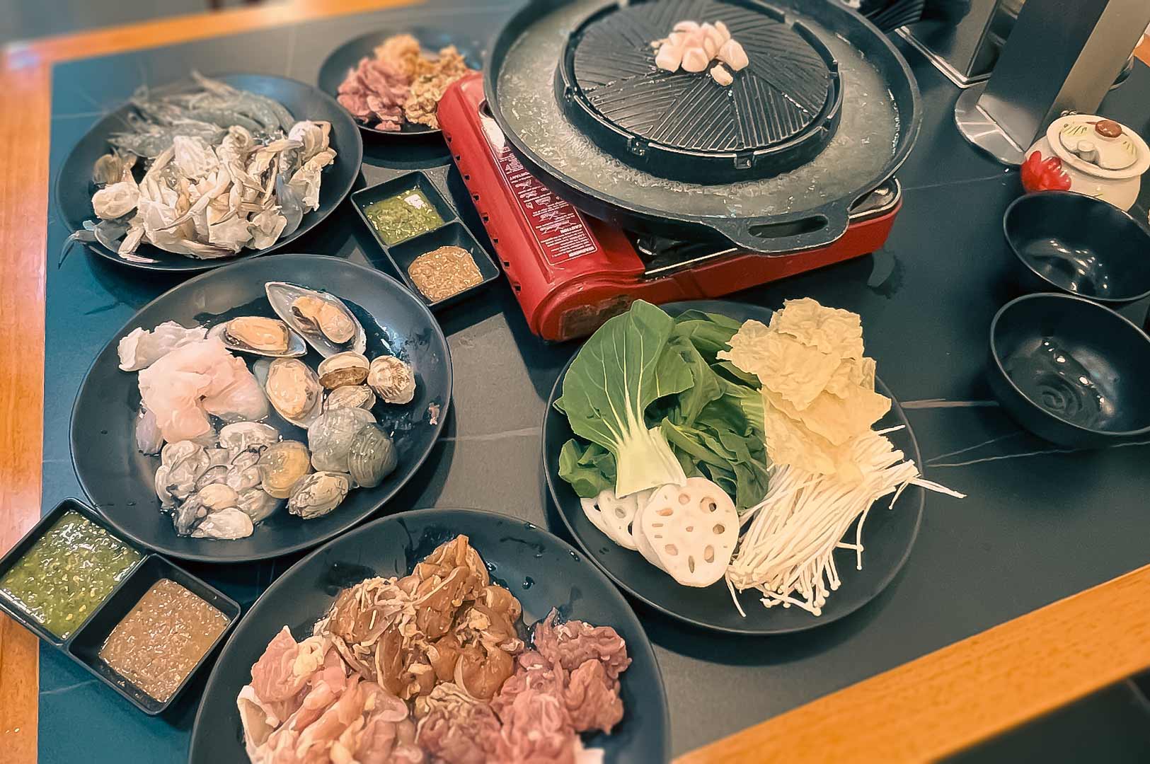 Spread of fresh Mookata ingredients including seafood, sliced meats, vegetables, and dipping sauces around a tabletop grill.