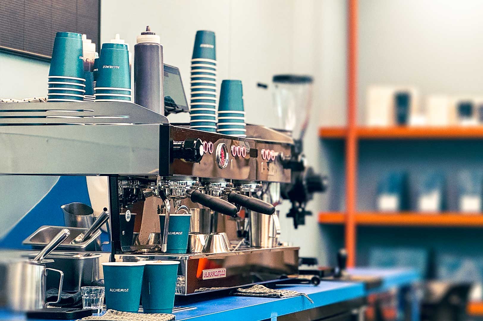 Espresso machine setup at Alchemist café with stacked teal coffee cups, brewing tools, and grinders on a blue counter.