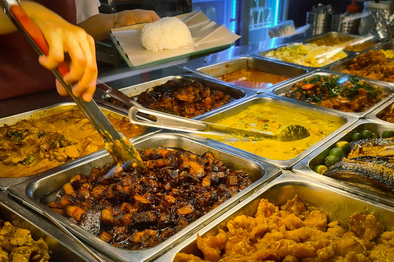 Casual dining moment at food stall where vendor serves customer selecting meals from buffet-style trays of diverse dishes.
