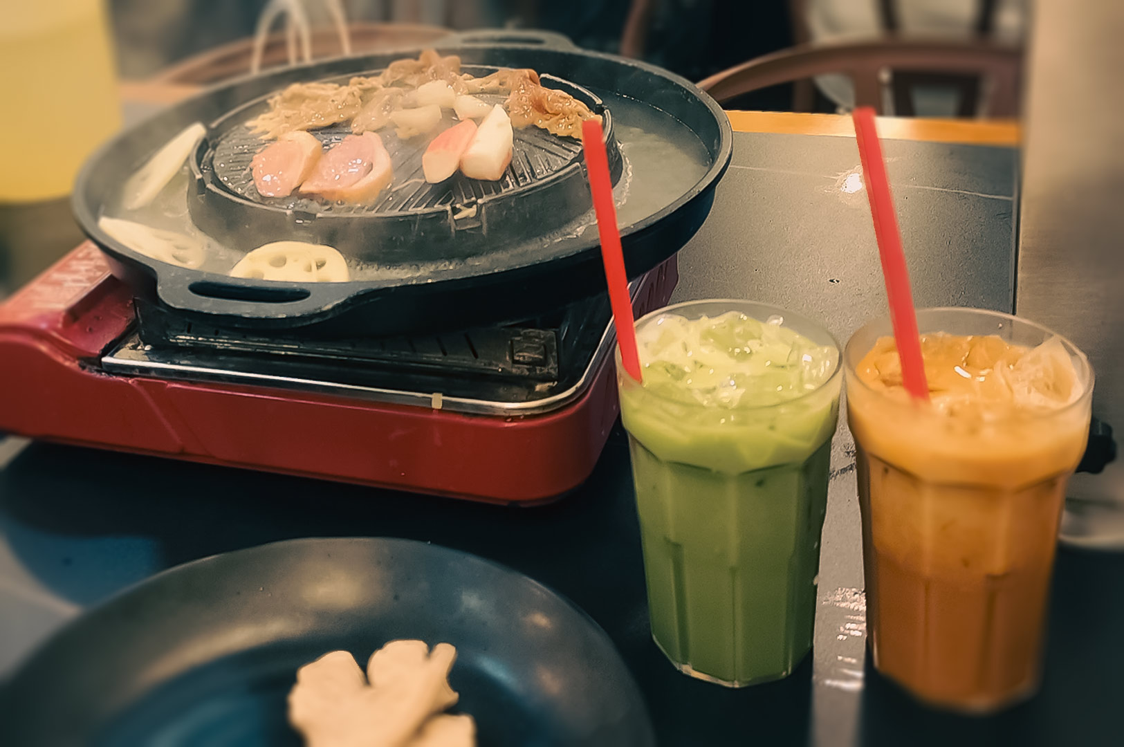 Mookata grill cooking meat and seafood beside iced Thai milk tea and iced green tea.