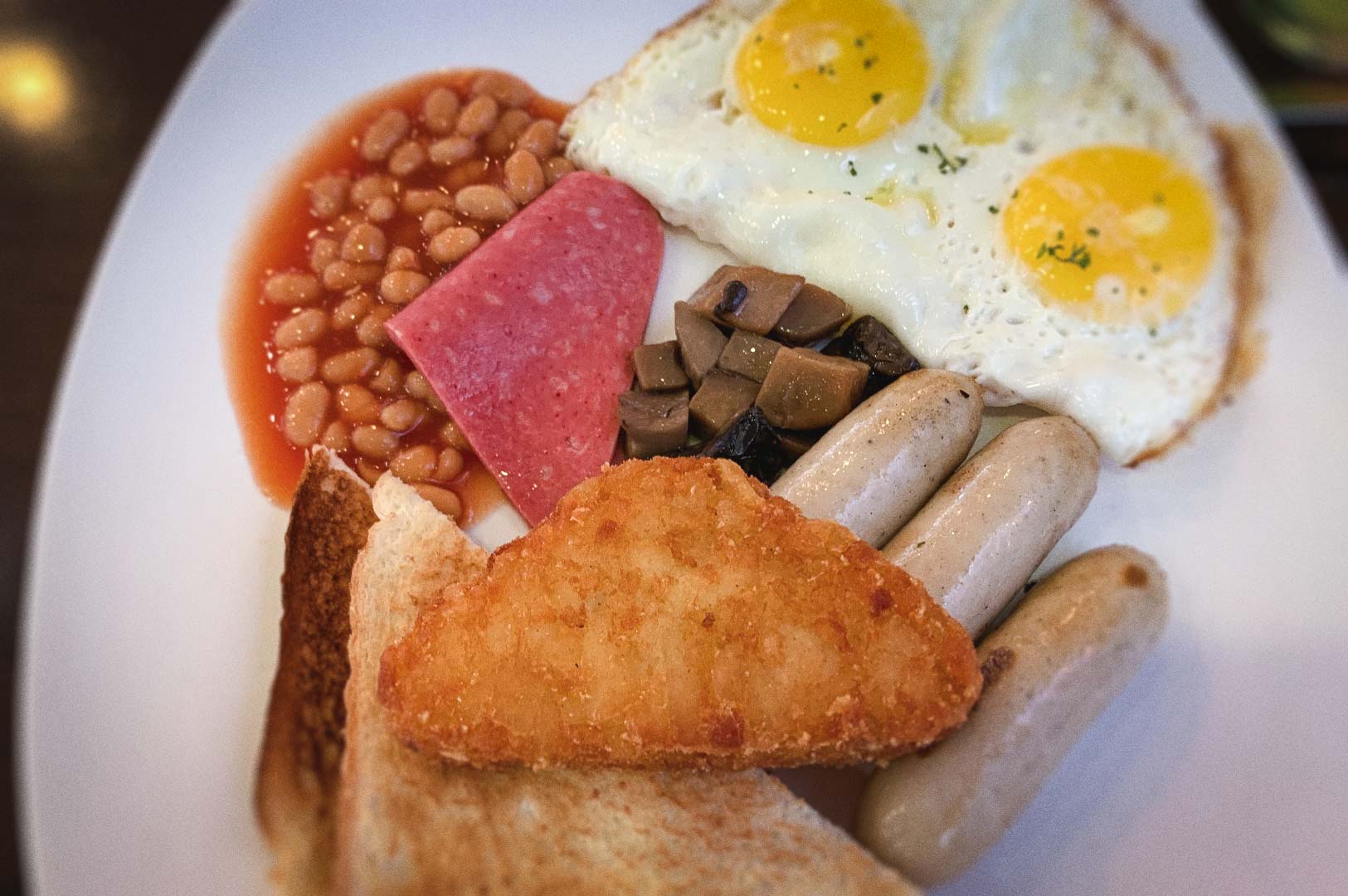 Breakfast plate with sunny‑side‑up eggs, baked beans, sliced ham, sautéed mushrooms, sausages, toast, and a crispy hash brown.