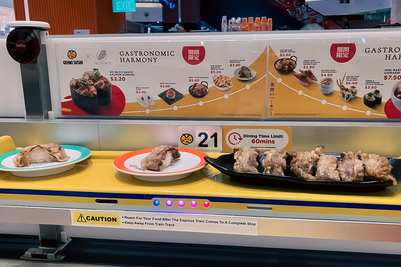 Sushi plates traveling on the Genki Sushi conveyor belt in front of a menu display featuring limited‑edition items.