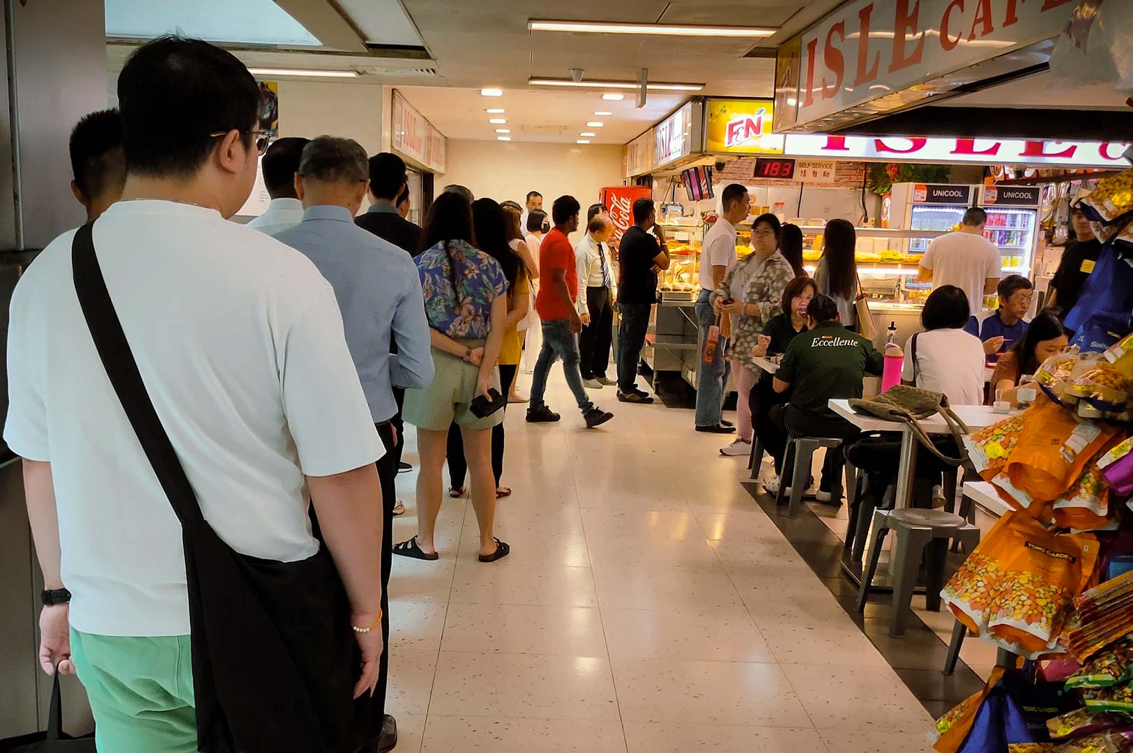 Customers queue up to order at busy food stall with display of cooked dishes.