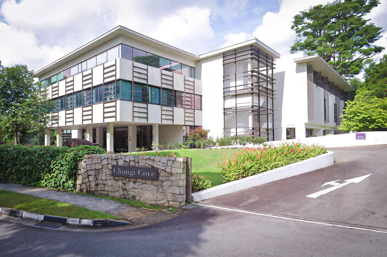 Exterior view of Changi Cove, featuring modern white buildings with large windows, landscaped greenery, and a stone entrance sign along a curved driveway.