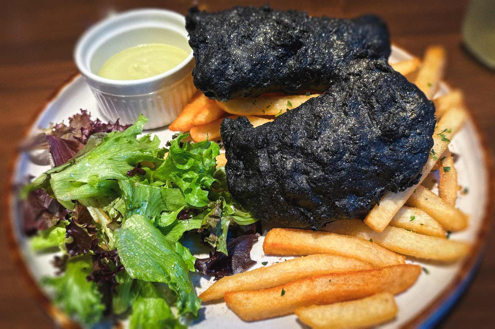 Plate of charcoal‑battered fish served with golden fries, mixed green salad, and a small cup of creamy dipping sauce.