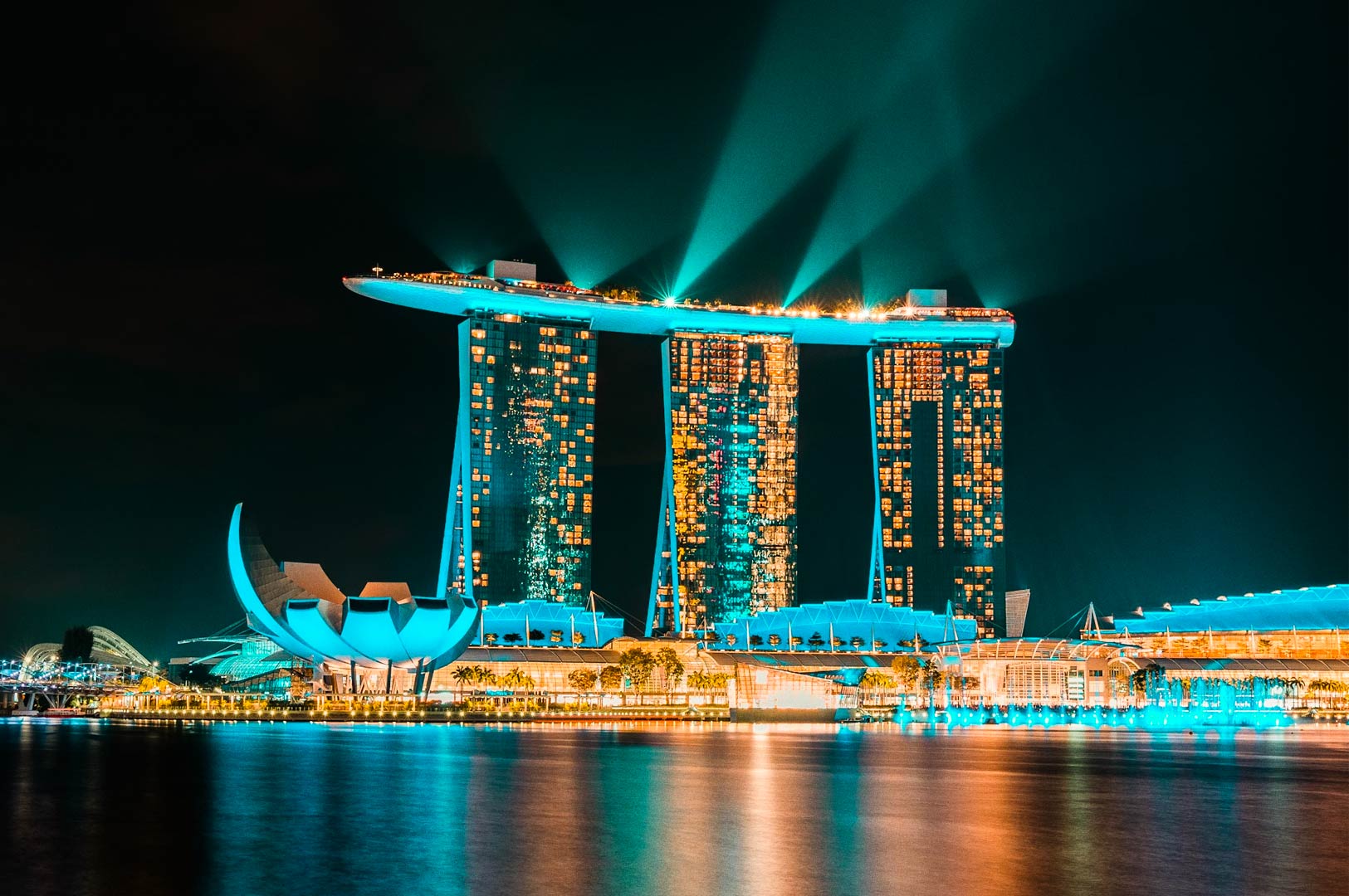 Night view of a waterfront resort with illuminated towers and sky beams.