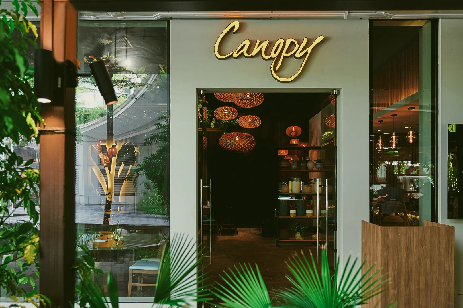 Wide-angle eye-level shot of Canopy restaurant entrance at Changi City Point, featuring green plants, glass façade, illuminated signage, and a welcoming modern café exterior.