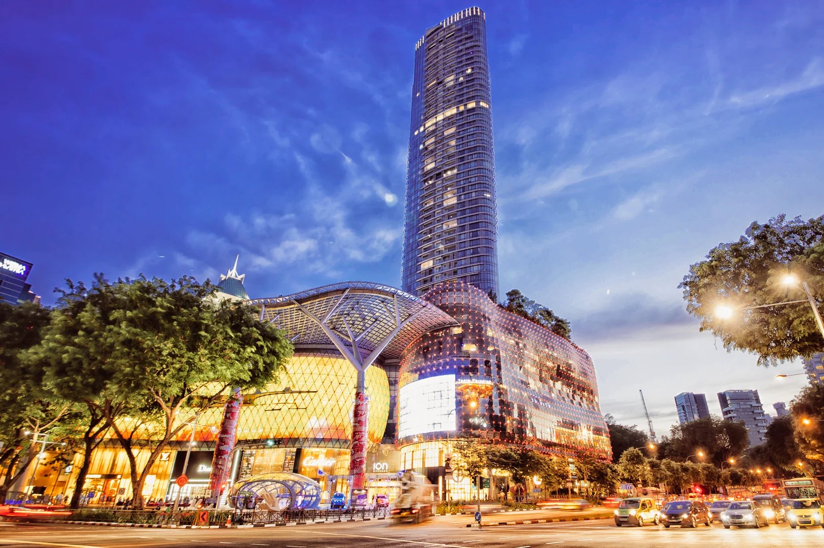 1 ION Orchard Cafes Low‑angle exterior shot of ION Orchard shopping mall at dusk, highlighting modern glass architecture, illuminated facade, surrounding trees, and city traffic.