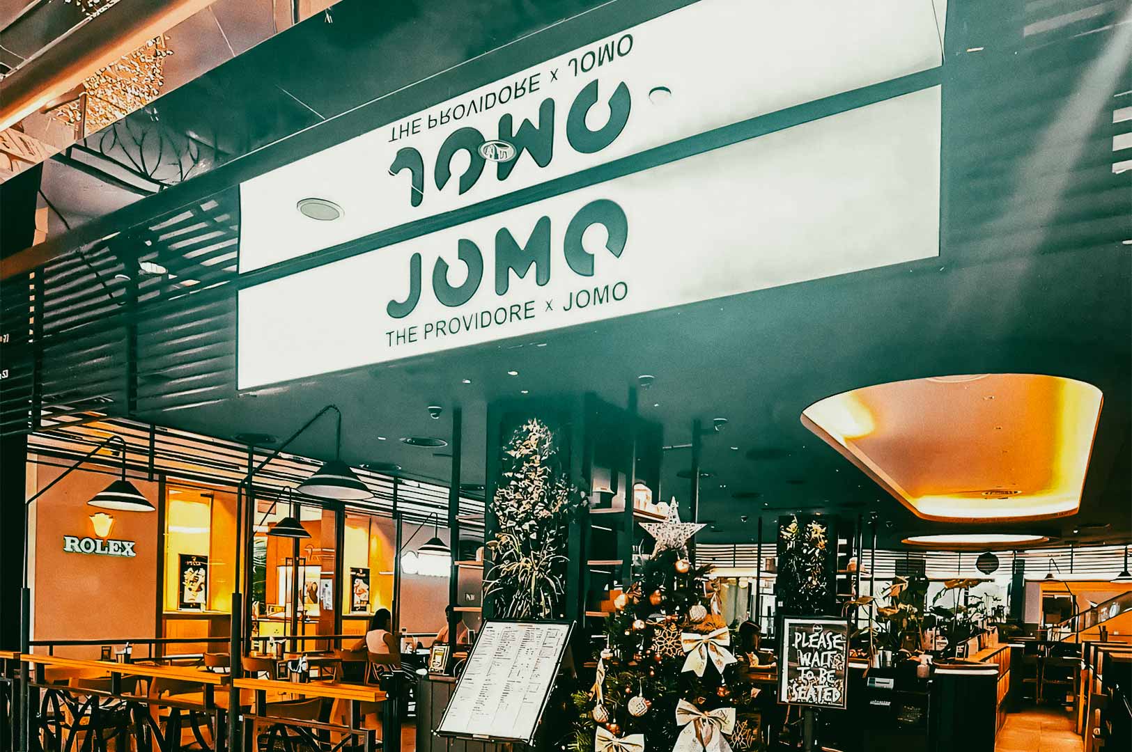 The exterior of a restaurant named ‘JOMO – The Providore x JOMO,’ with seating visible inside and Christmas decorations, including a decorated tree, displayed at the entrance.