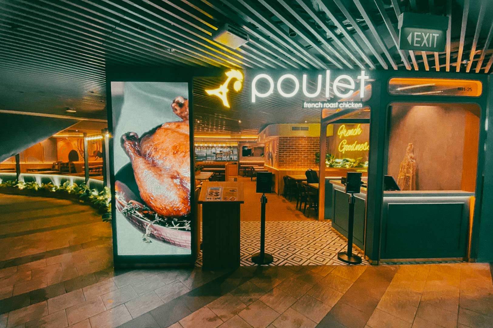 Interior of a Poulet restaurant with customers seated at wooden tables, decorated with wall art, indoor plants, and a neon sign reading ‘French Goodness.
