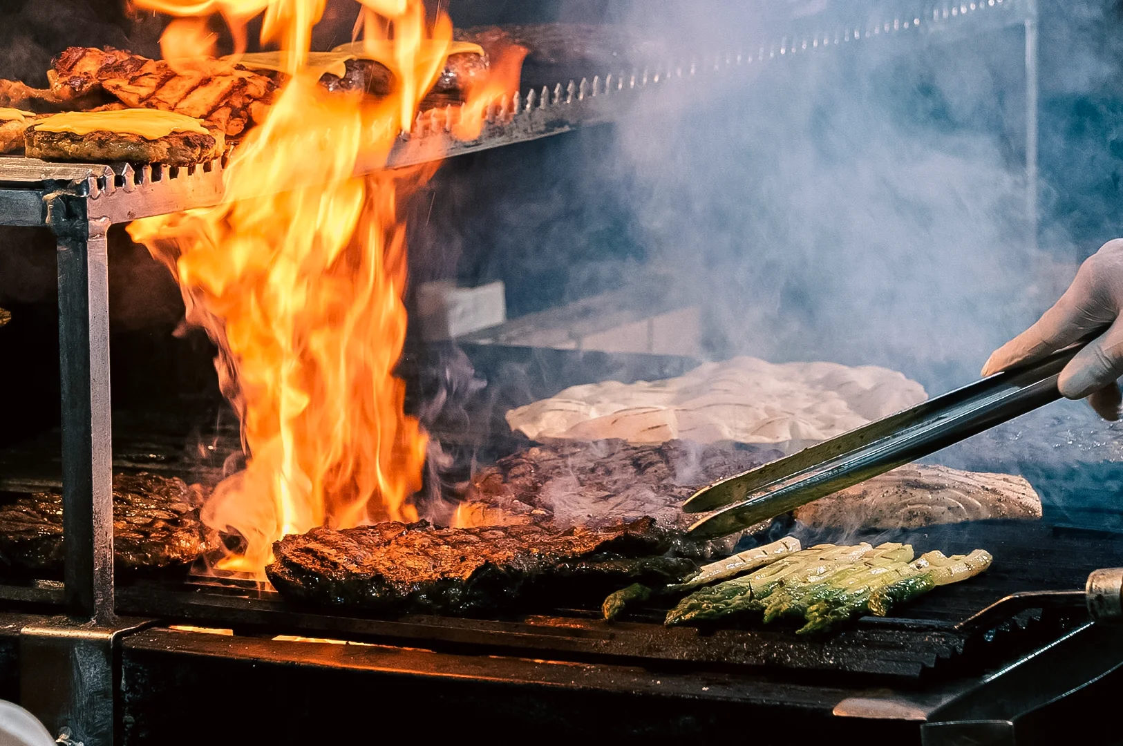 Close‑up action shot of steaks and vegetables grilling over open flames on a commercial restaurant grill, with metal tongs turning food amid rising smoke, highlighting high‑heat cooking, chef techniques, and premium steakhouse preparation.