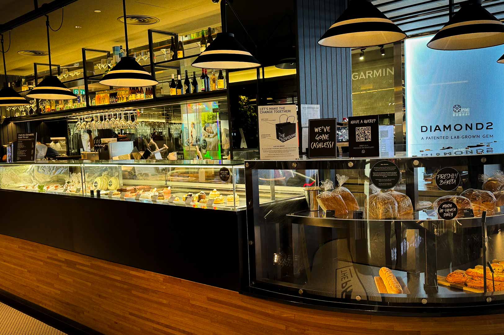 A bakery and café counter displaying assorted pastries and loaves of bread behind glass, with hanging black pendant lights above and shelves of bottles and glassware in the background.