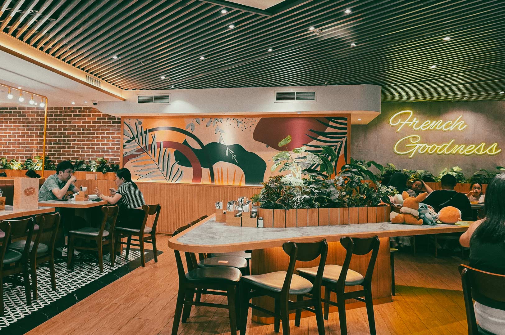 Interior of a Poulet restaurant with customers seated at wooden tables, decorated with wall art, indoor plants, and a neon sign reading ‘French Goodness.