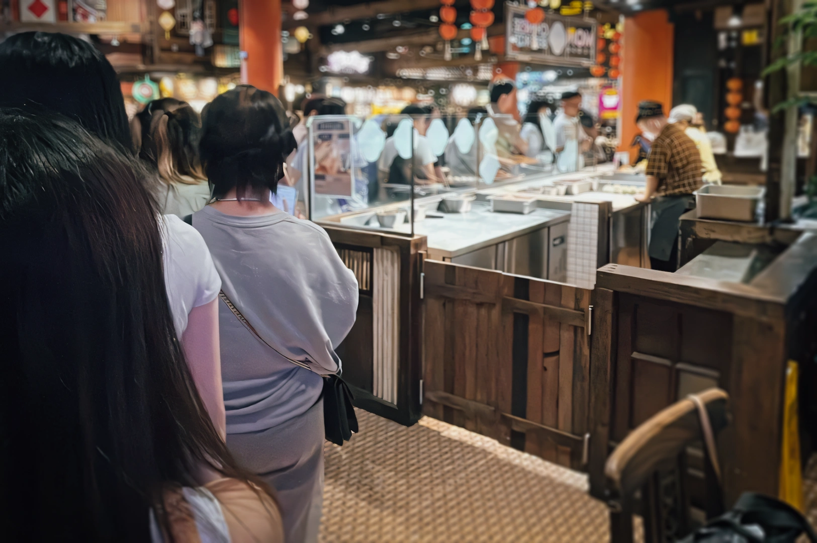 Eye‑level wide shot of a busy indoor food hall with customers lining up at open kitchen counters, wooden stall fronts, and warm ambient lighting, highlighting modern food court dining and popular Asian cuisine stalls.