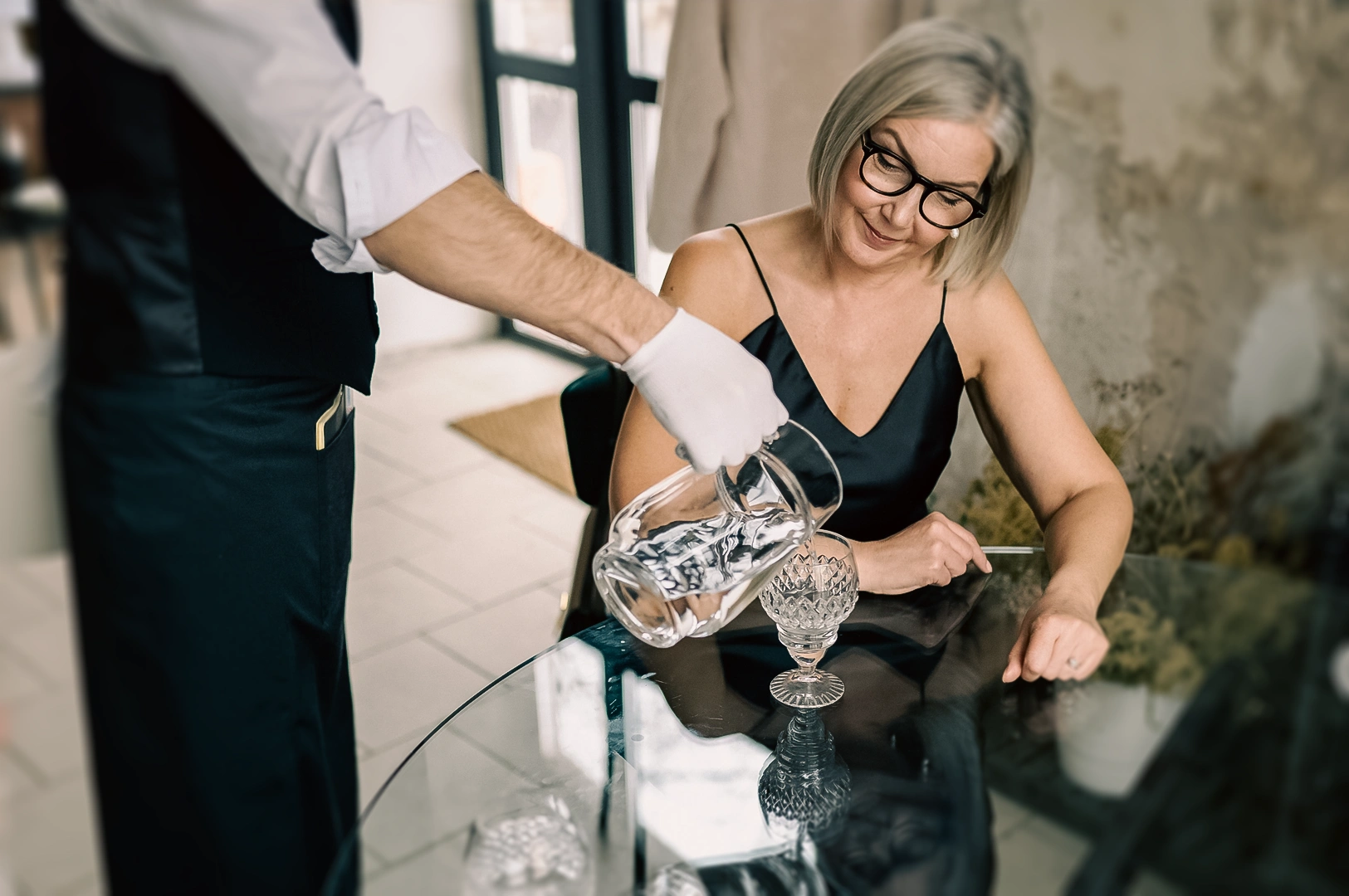 Eye‑level medium shot of table‑side water service as a server pours water into a crystal glass at a modern restaurant table, emphasizing fine dining experience, attentive hospitality, and elegant dining atmosphere.