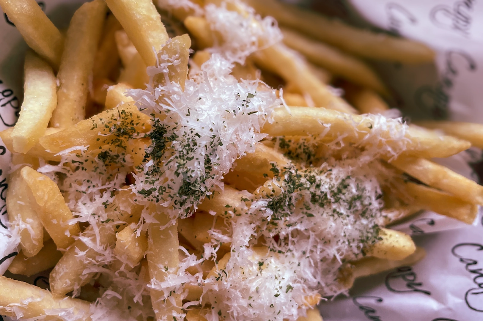 Close-up overhead shot of golden French fries topped with finely grated cheese and herbs, served on branded paper lining, highlighting gourmet truffle fries presentation.