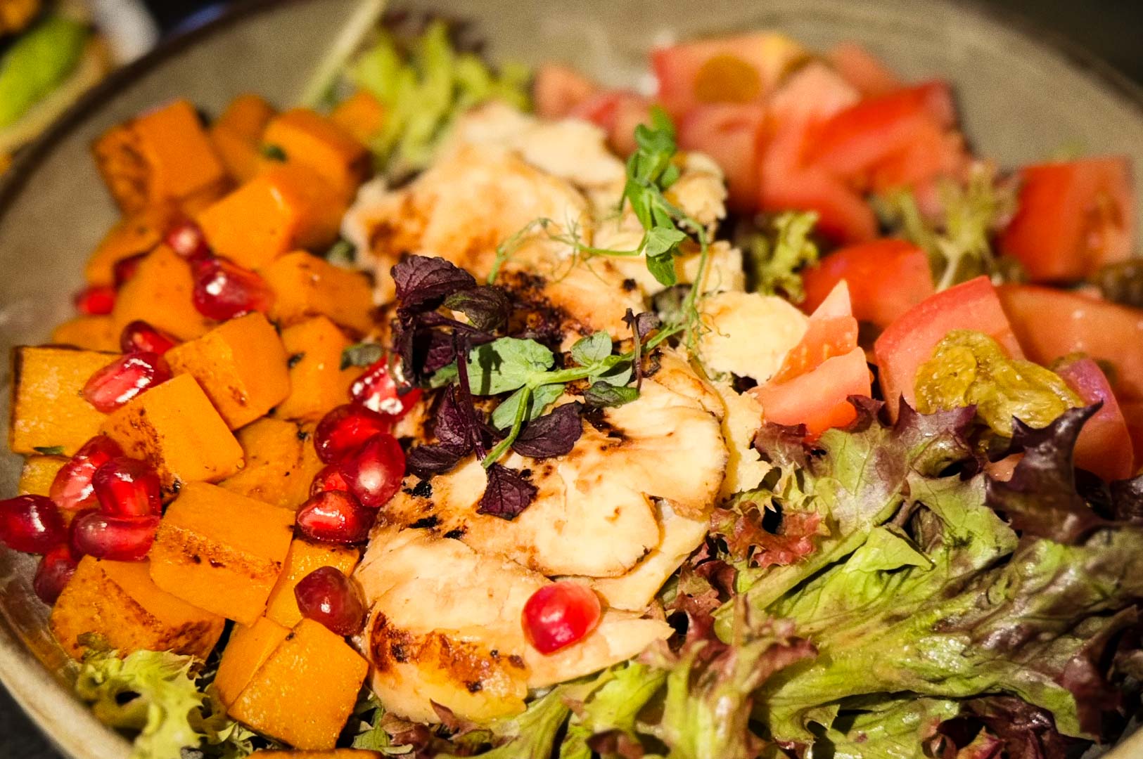 A colorful salad bowl featuring grilled chicken pieces, leafy greens, diced roasted pumpkin, fresh tomatoes, and pomegranate seeds, topped with small herbs.