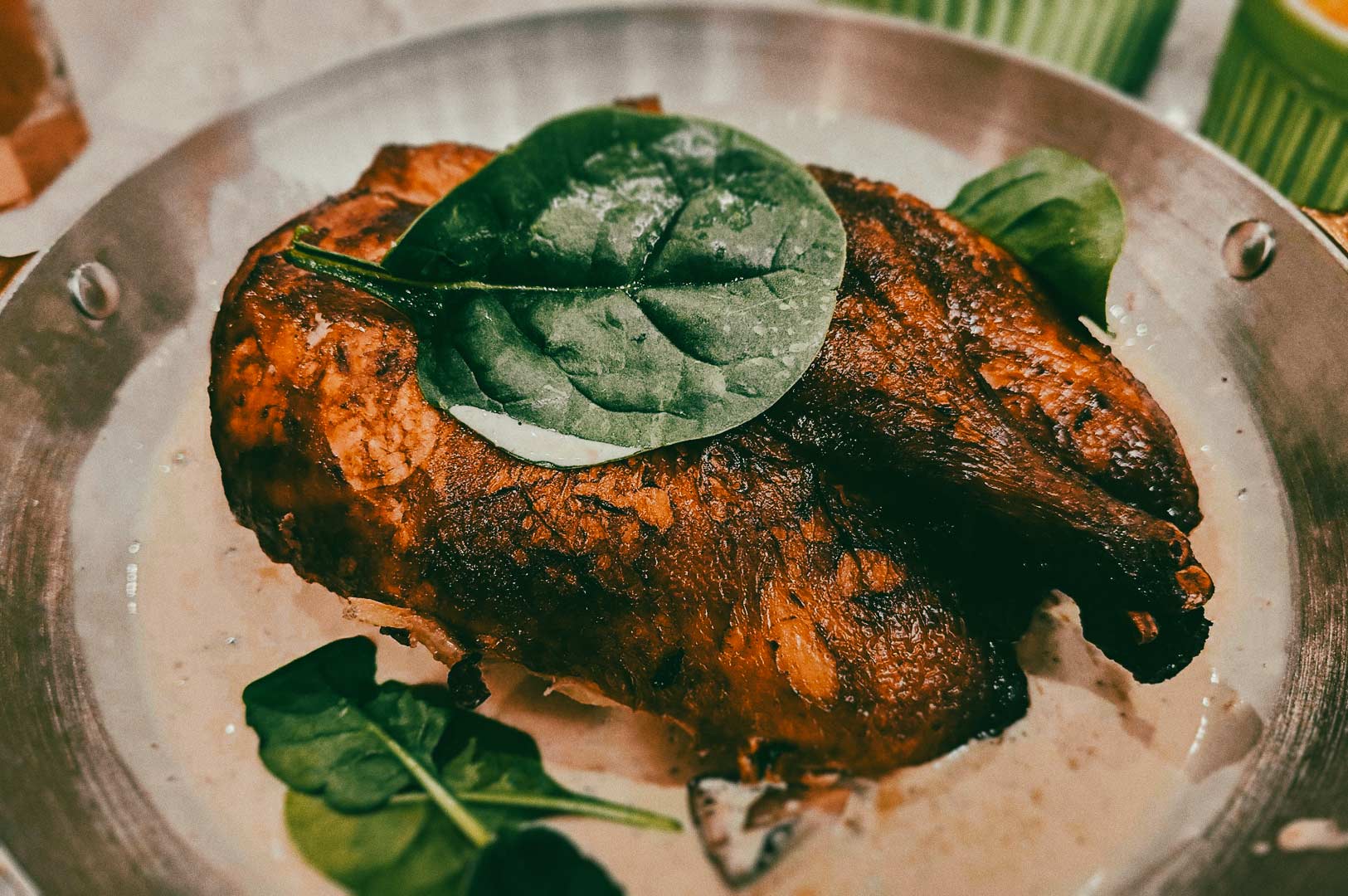 A roasted chicken served in a round metal pan, topped with fresh spinach leaves and surrounded by a creamy sauce.