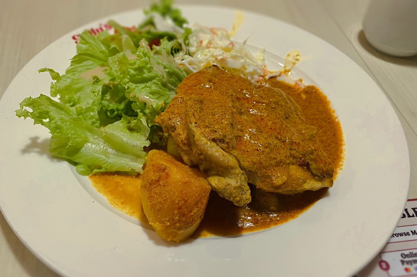 Close-up overhead shot of a plated curry chicken dish served with potatoes, lettuce, and shredded vegetables on a white plate, highlighting rich sauce texture and casual restaurant presentation.