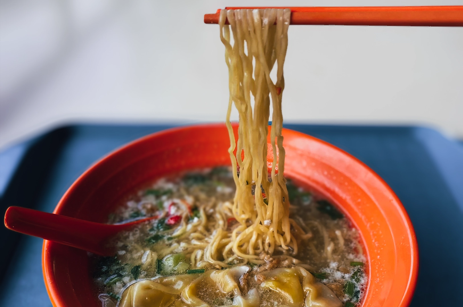 Close‑up overhead shot of bak chor mee with springy egg noodles lifted by chopsticks from a bowl of clear broth, featuring minced pork, dumplings, and garnishes, highlighting classic Singapore hawker noodles and local comfort food.