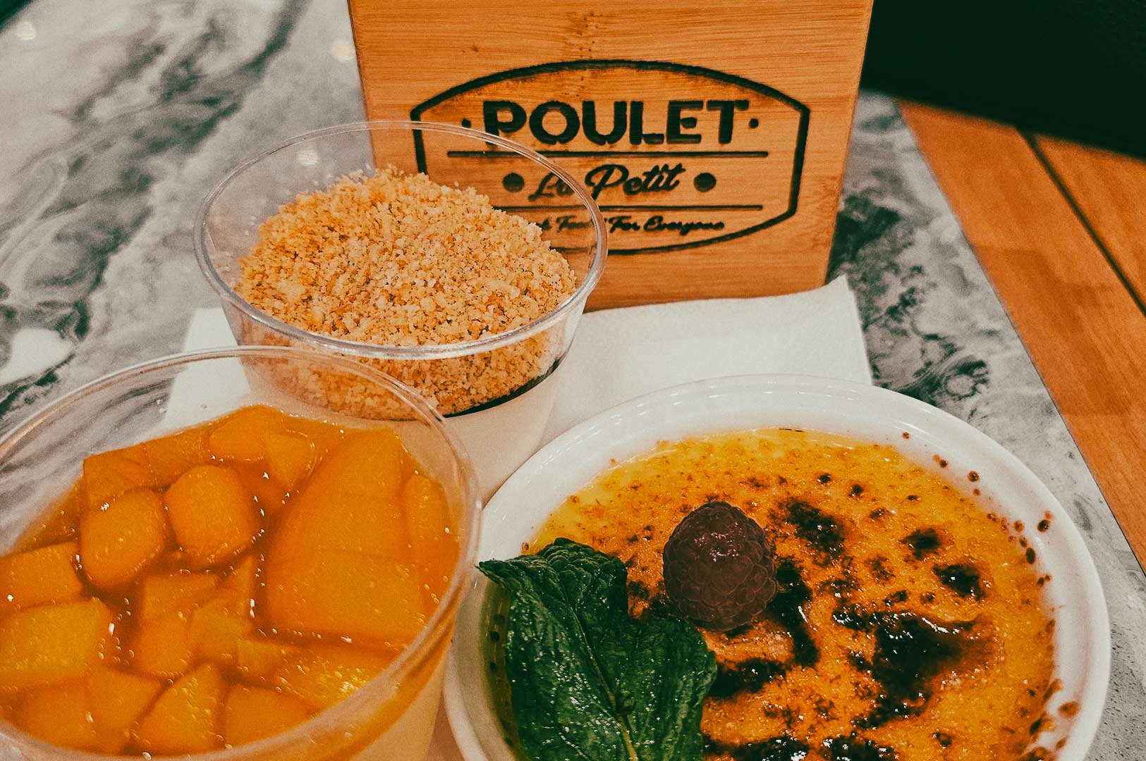 Three dessert items displayed on a table, including a cup of diced mango, a cup of crumb topping, and a crème brûlée garnished with mint, with a wooden Poulet-branded box in the background.