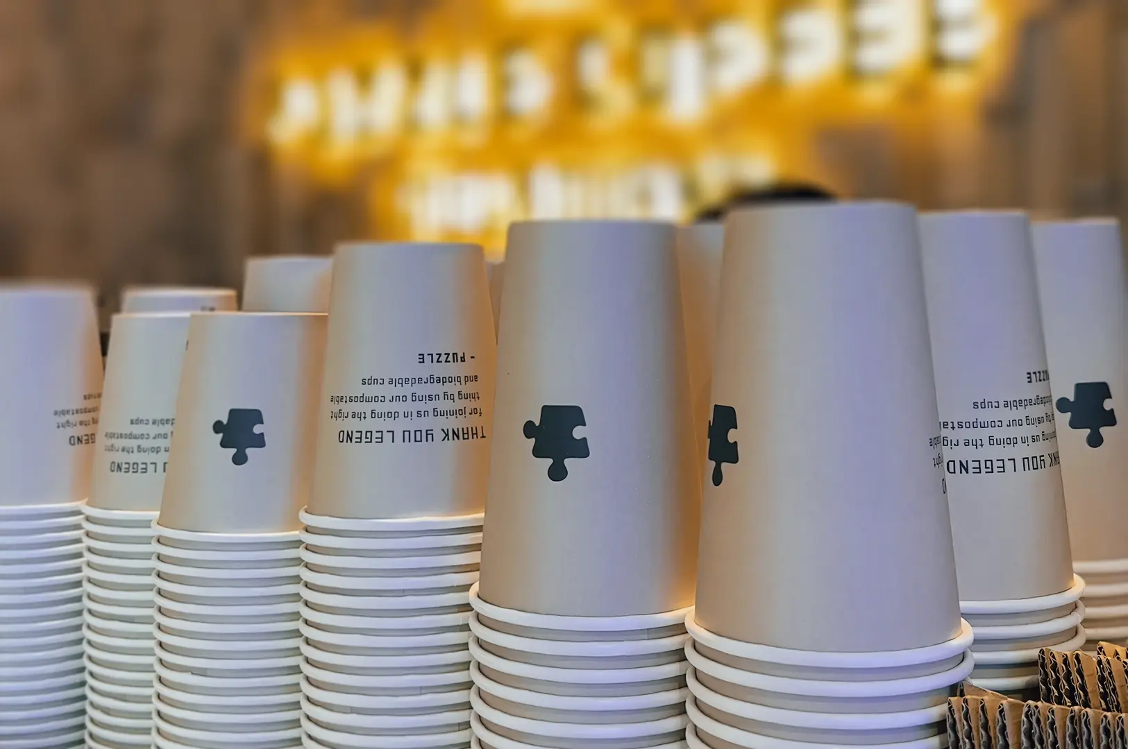 Low-angle close-up shot of stacked Puzzle Coffee takeaway cups with logo branding, arranged behind the counter with warm café lighting in the background.
