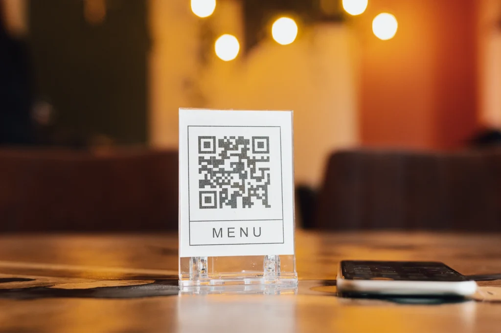 Shallow depth-of-field eye-level shot of a tabletop QR code menu displayed in a clear acrylic stand labeled “Menu,” placed on a restaurant table with warm ambient lighting and a smartphone beside it, highlighting digital dining and contactless menu trends.