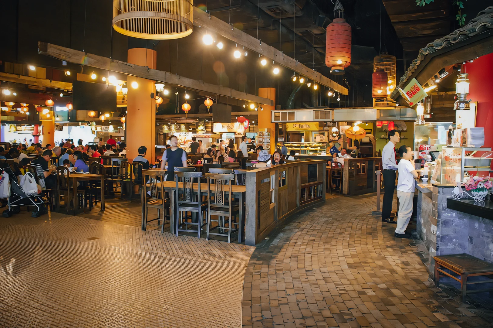 Wide-angle interior shot of a busy Asian mall food court with diners seated at communal wooden tables, warm ambient lighting, hanging lanterns, and multiple food stalls creating a lively marketplace atmosphere.