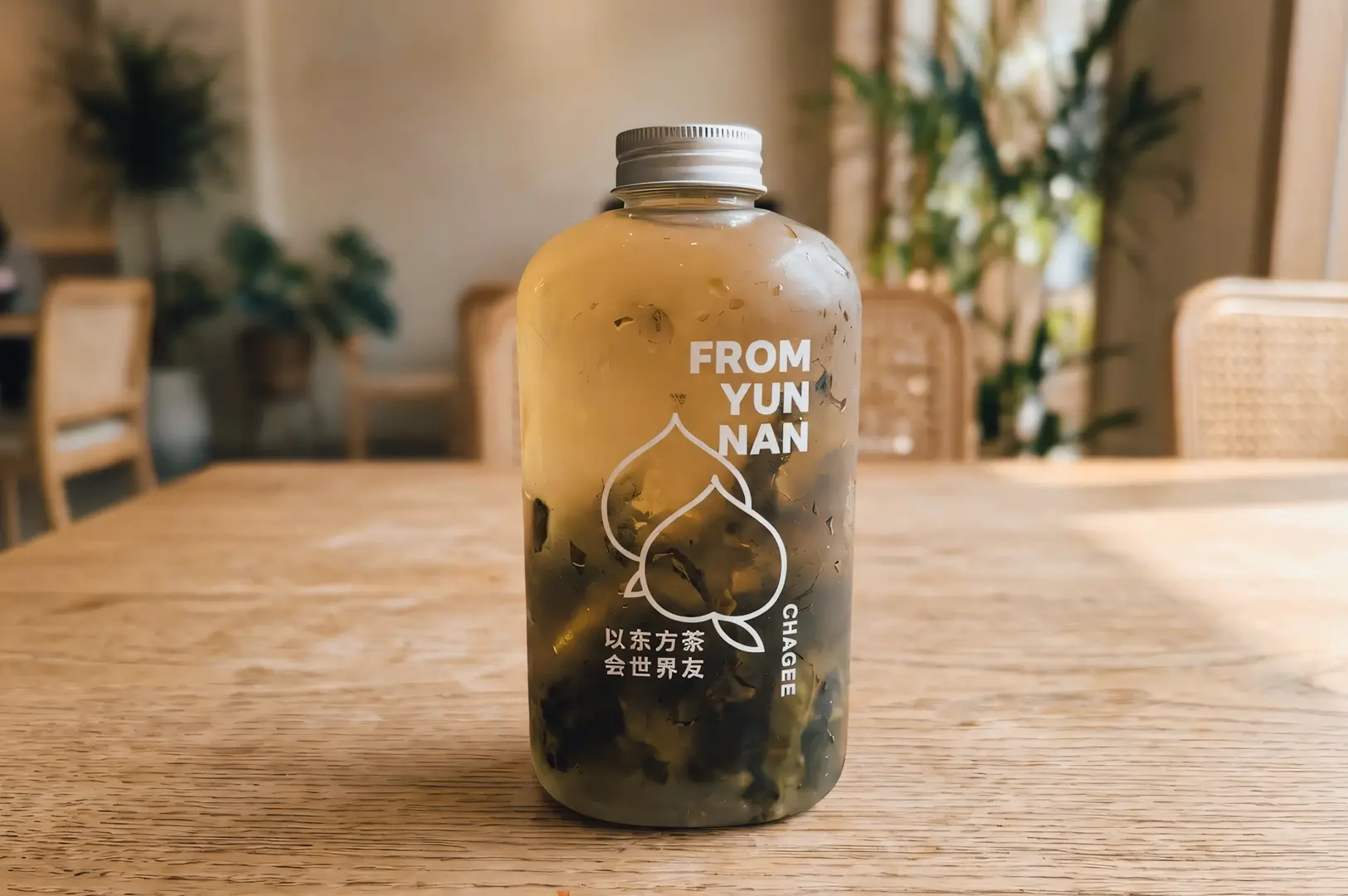 Close‑up product shot of a CHAGEE Cold Brew Tie Guan Yin tea bottle, photographed at eye level on a wooden table, showing layered tea tones and visible tea leaves through the glass with soft natural café lighting in the background.
