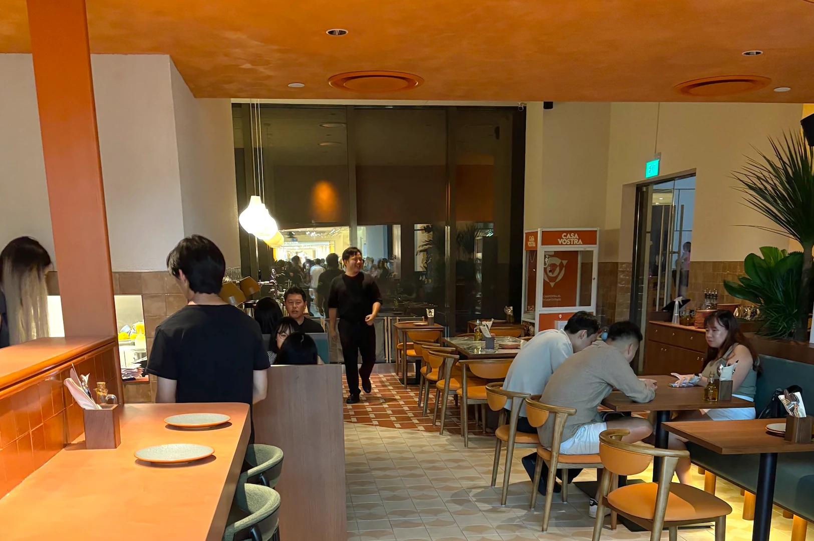 Wide-angle interior shot of Casa Vostra restaurant with diners seated at wooden tables, warm ambient lighting, tiled floors, and an open dining layout creating a lively Italian bistro atmosphere.