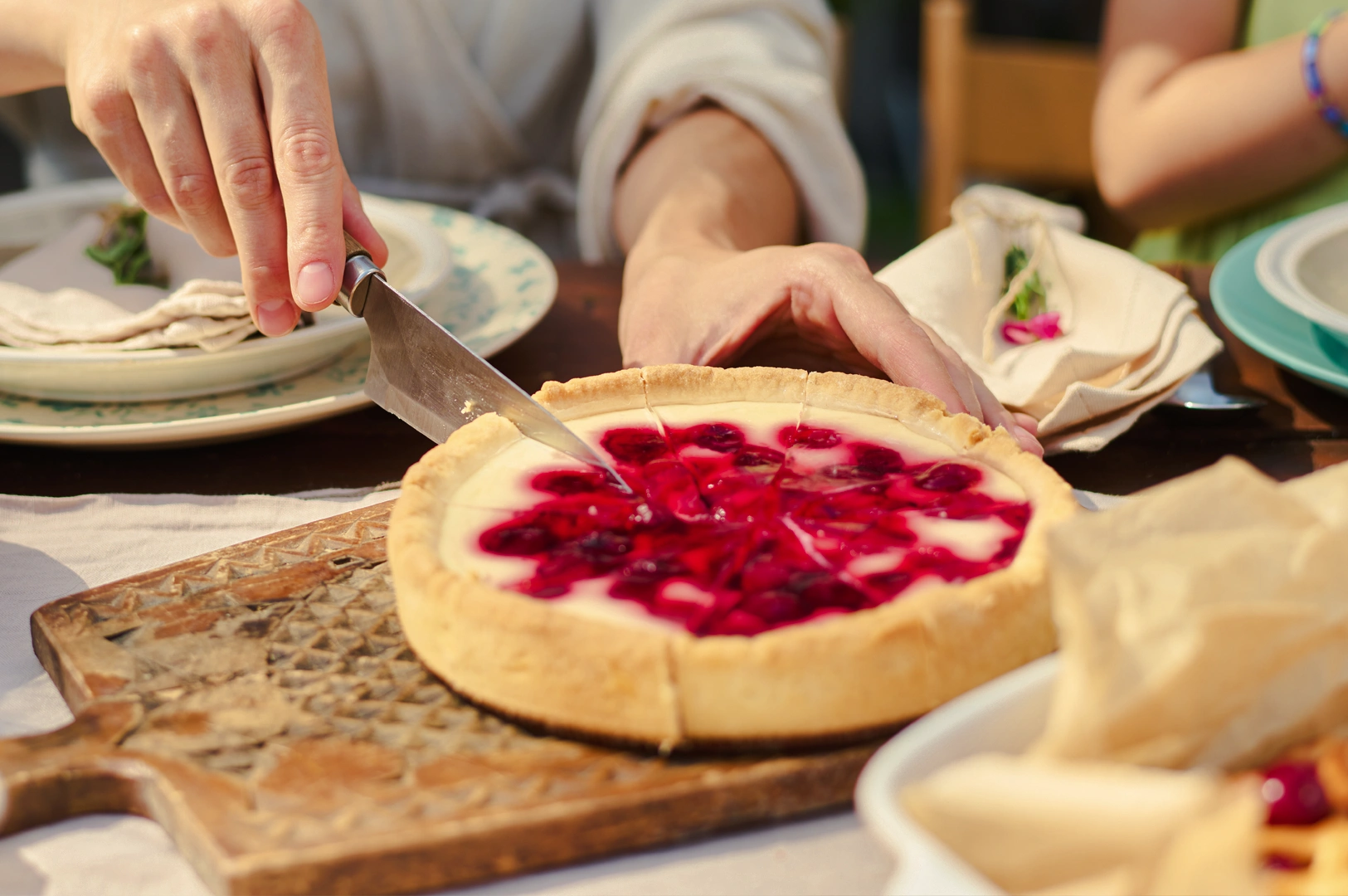 2 Limited-Time Menu Close‑up eye‑level shot of hands slicing a cherry‑topped cheesecake on a wooden serving board, with plates and tableware arranged around the dessert in a casual dining setting.