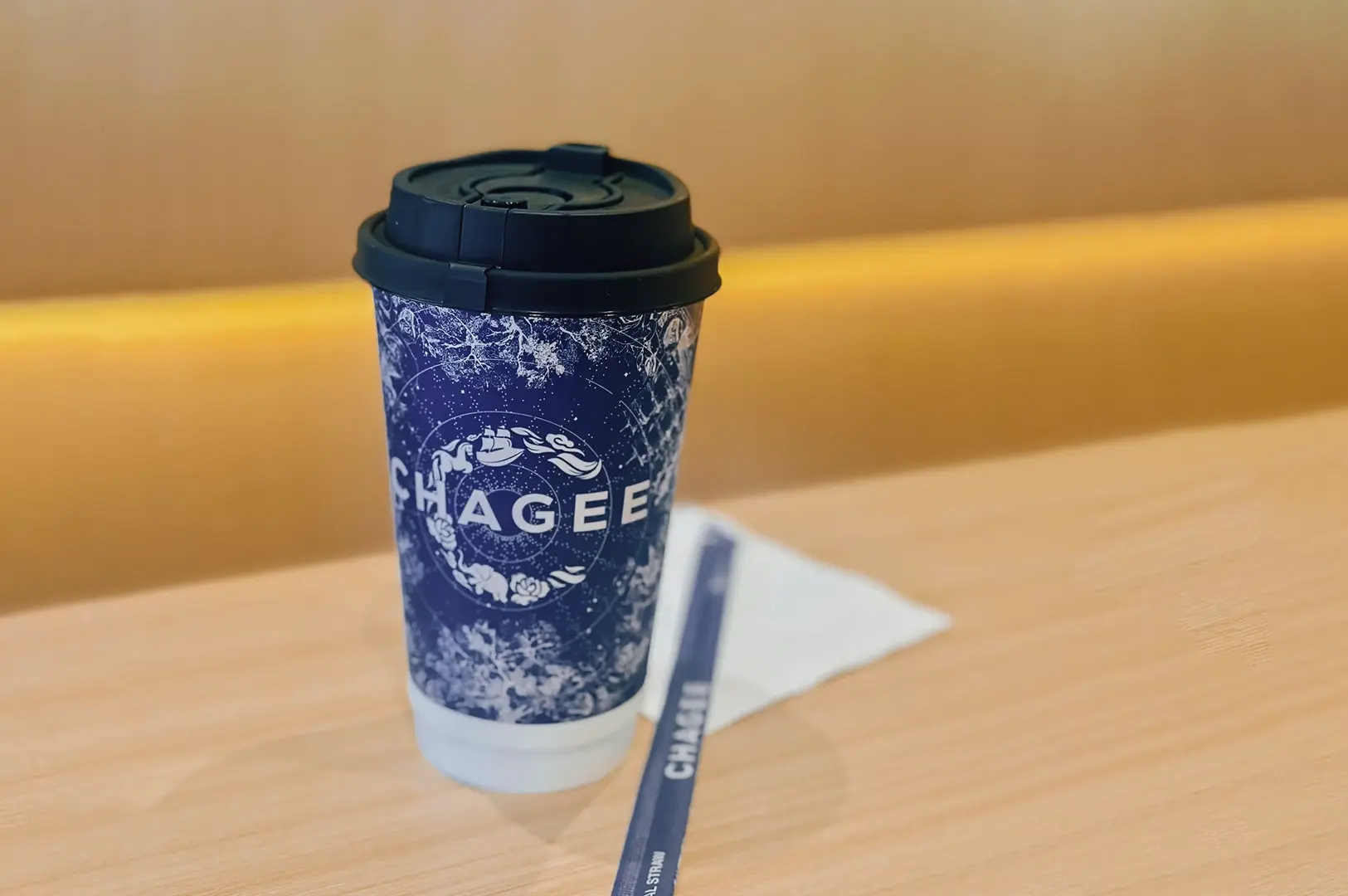 Close‑up product shot of a CHAGEE branded takeaway tea cup with lid on a wooden table, photographed from a slightly elevated angle with shallow depth of field, highlighting the navy floral logo and minimalist café setting.