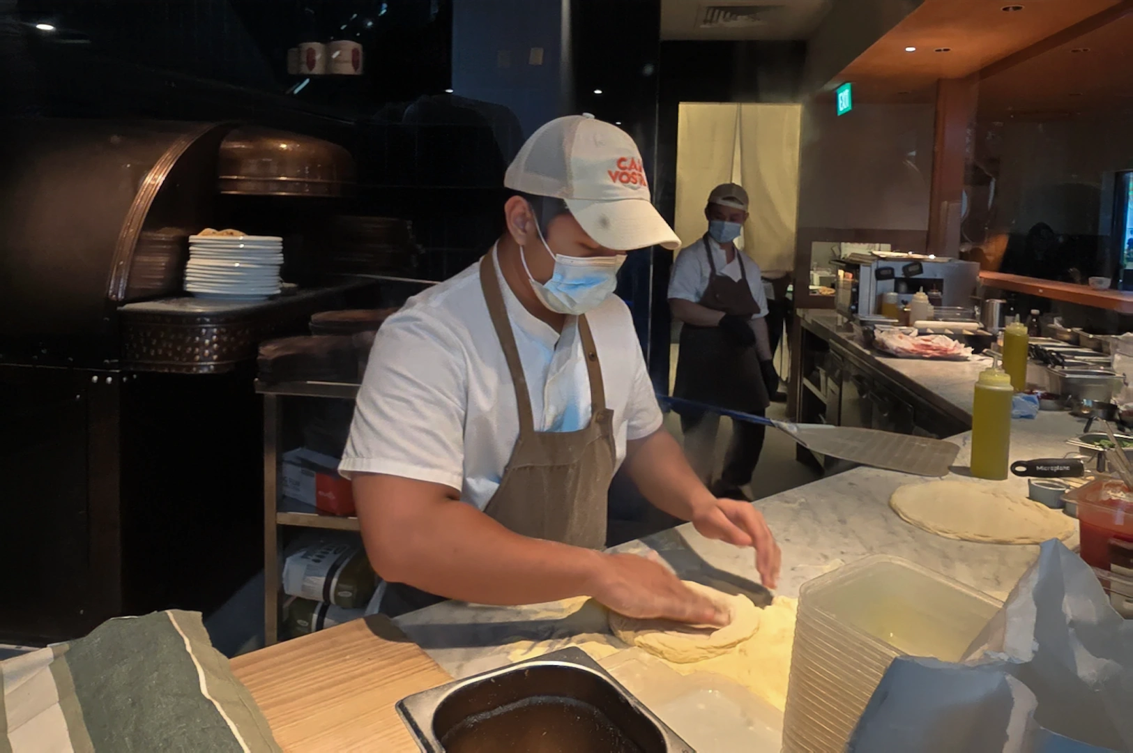 Medium close-up eye-level shot of a Casa Vostra kitchen staff member hand-stretching fresh pizza dough on a marble counter in an open kitchen, showcasing artisanal Italian pizza preparation at JEM mall.