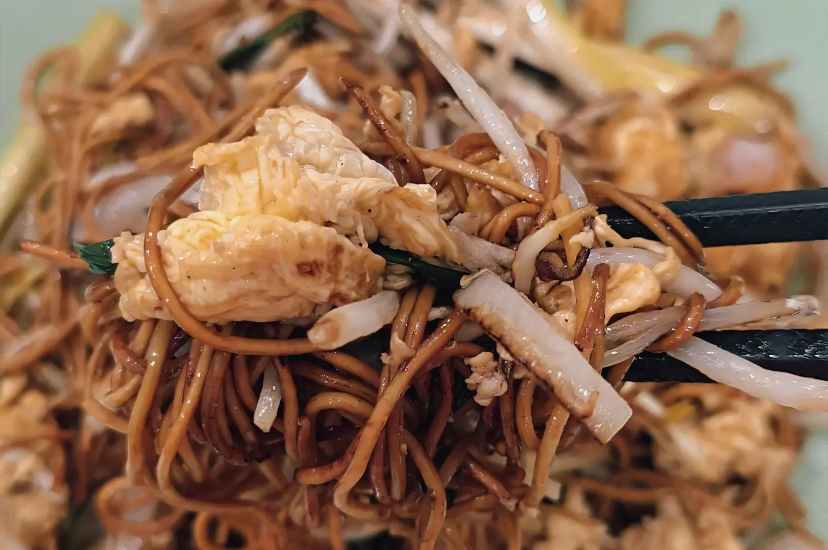 Extreme close-up, eye-level shot of Hong Kong–style stir-fried noodles with egg, bean sprouts, and scallions being lifted with chopsticks, highlighting glossy texture and freshly cooked wok-fried noodles.