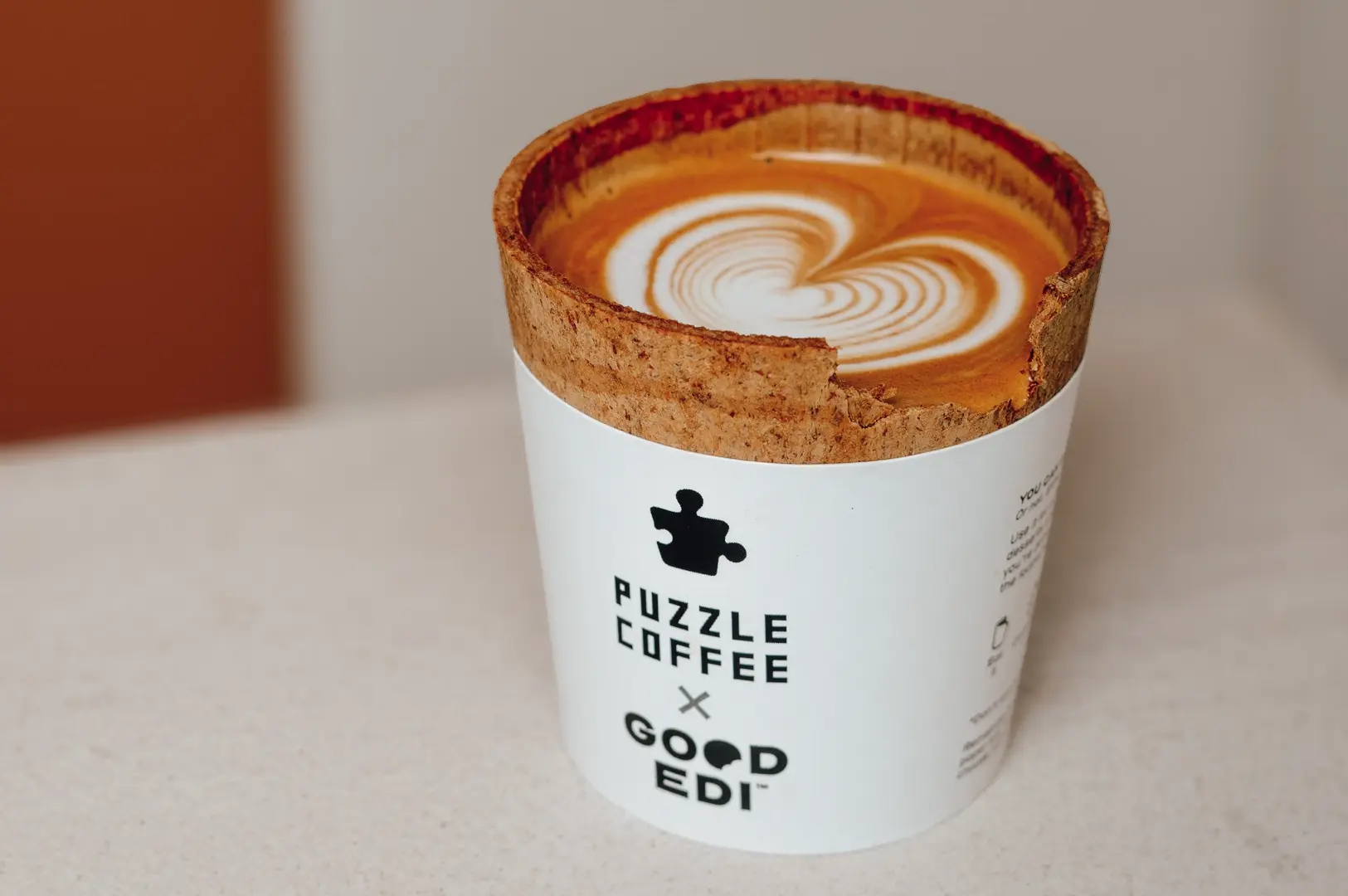 Close-up eye-level shot of a latte with heart-shaped latte art served in an edible cookie cup, branded Puzzle Coffee x Good Edi, placed on a minimalist café countertop.