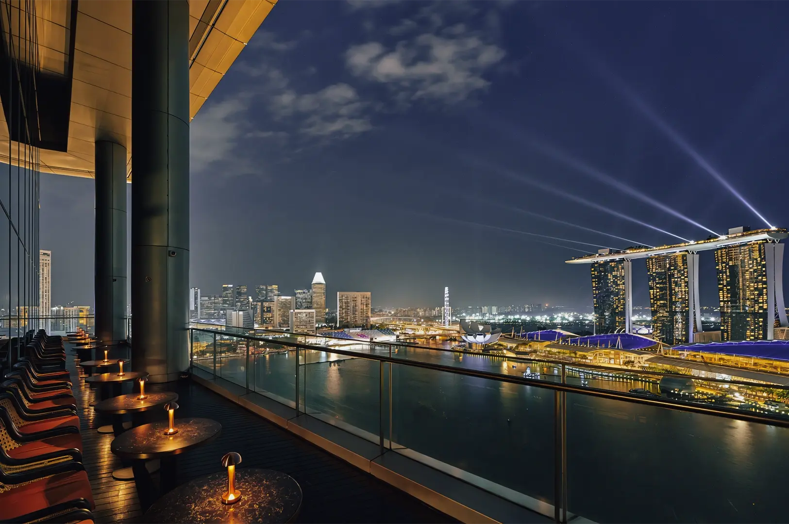 Rooftop lounge overlooking a nighttime city skyline and waterfront, with candle‑lit tables and glass railings, photographed from a balcony‑level wide shot capturing urban luxury and panoramic city views.