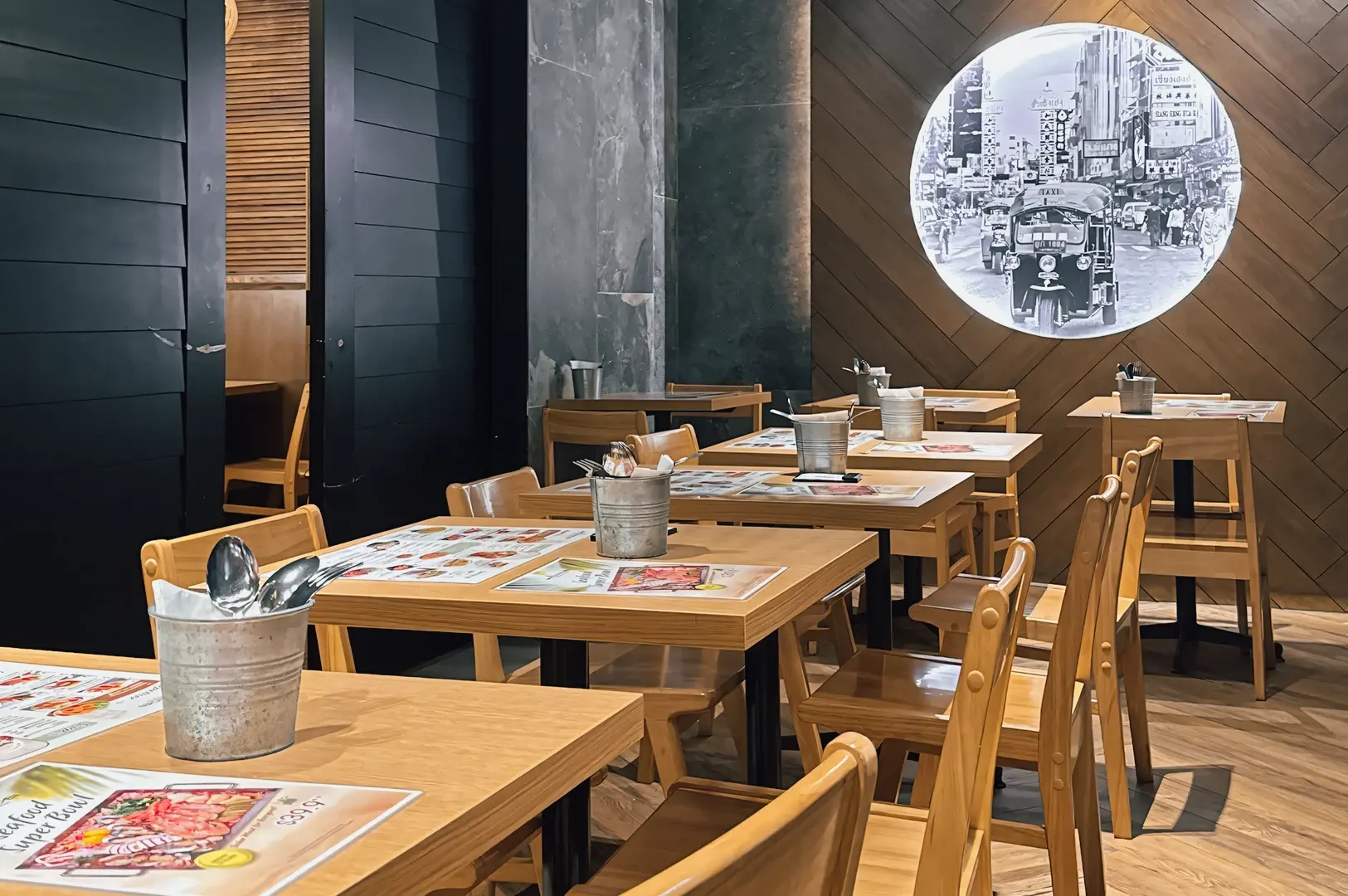 Wide‑angle interior shot of Sanook Kitchen restaurant dining area, photographed at eye level, featuring wooden tables and chairs, place settings with menus, warm lighting, and a circular black‑and‑white city photo mounted on a textured wall.